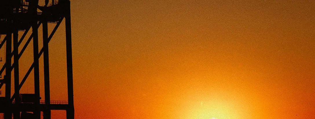 A silhouette of an industrial structure is contrasted against a vibrant sunset, with shades of orange and yellow blending into a warm sky, creating a serene atmosphere.