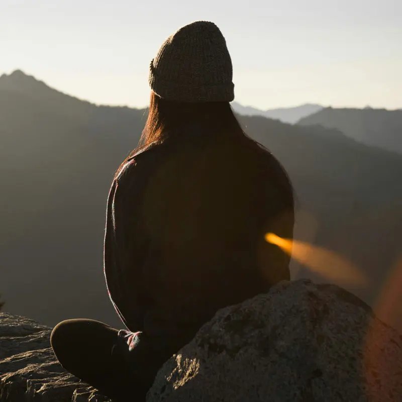 A person sits on a rocky outcrop, looking out over distant mountains at sunset. 