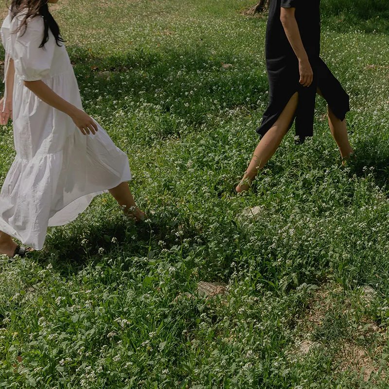 Two people walk through a lush green field dotted with white flowers. One is dressed in a flowing white dress, while the other wears a black outfit, both enjoying a sunny day outdoors.