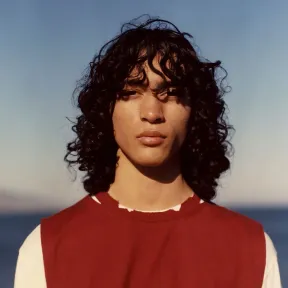 A young person with long, curly hair gazes confidently at the camera, wearing a red sleeveless top over a white shirt. The background features a blue sky and ocean.