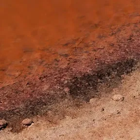The shoreline of a body of water with deep reddish hues, blending into a lighter sandy bank.